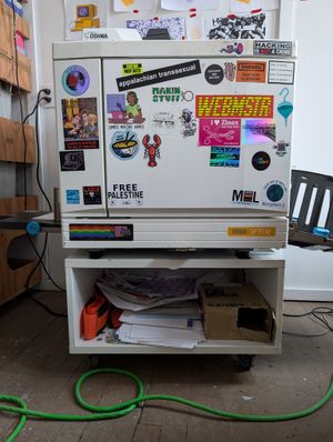 A riso printer covered in little stickers, resting on a wood floor with a green cable in front.