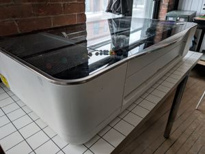 A photo of a laser cutter with a reflective top, on top of a table. The photo is taken at an angle.